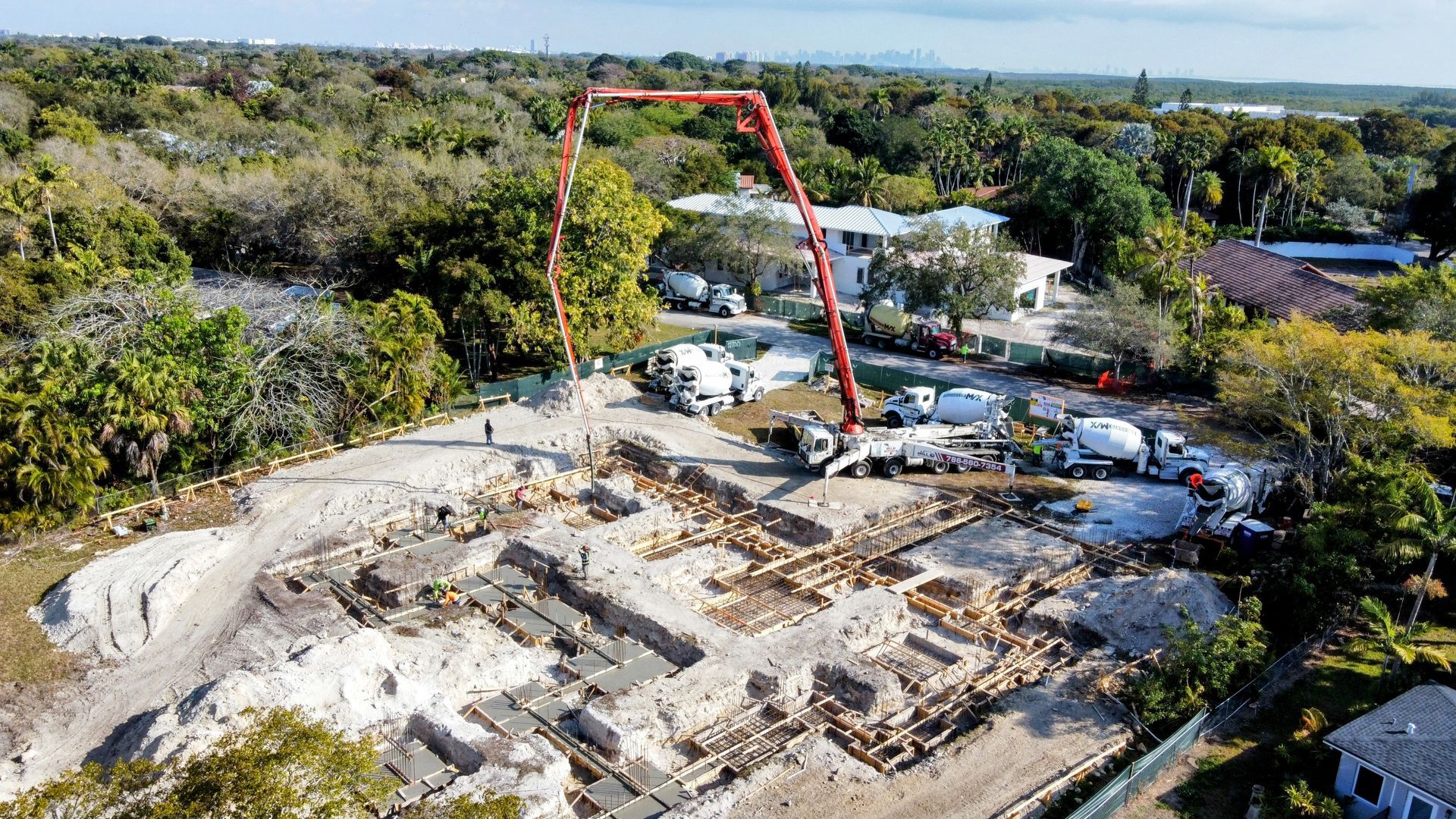 Construction site with concrete pump reaching over foundation. Several cement trucks and trees surround.