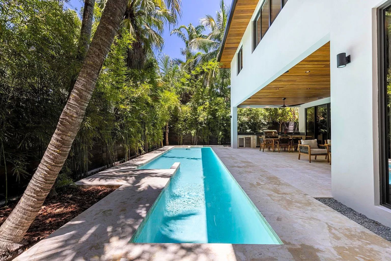 Swimming pool and patio of a modern house; blue water, light-colored walls, trees.
