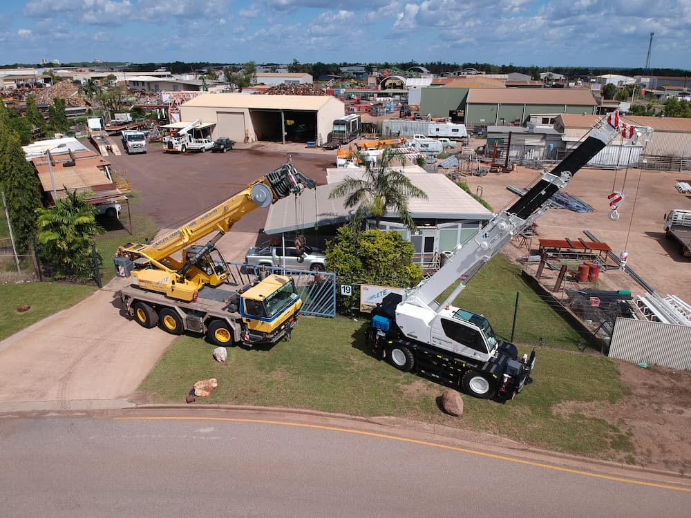 Two Cranes On A Lawn In Front Of Buildings, One Yellow, One White With Black Stripes, Ready To Lift