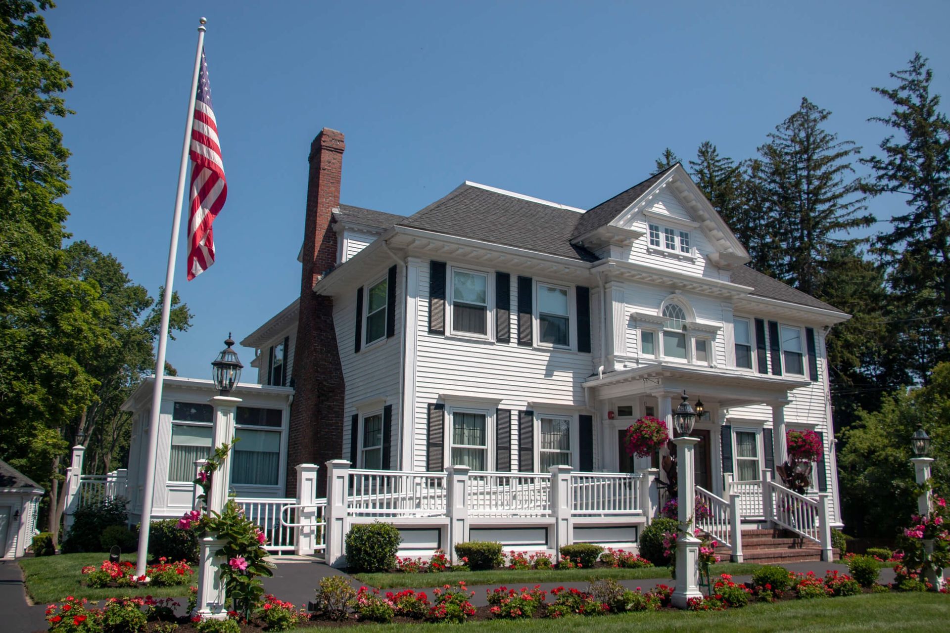 White house with black shutters, American flag, front porch, and red flowers.