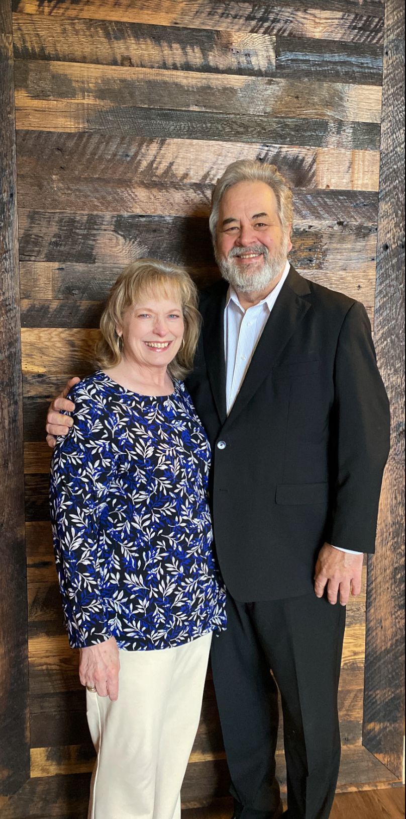 Couple posing in front of a wood wall. The woman is wearing a floral top, man in a suit.