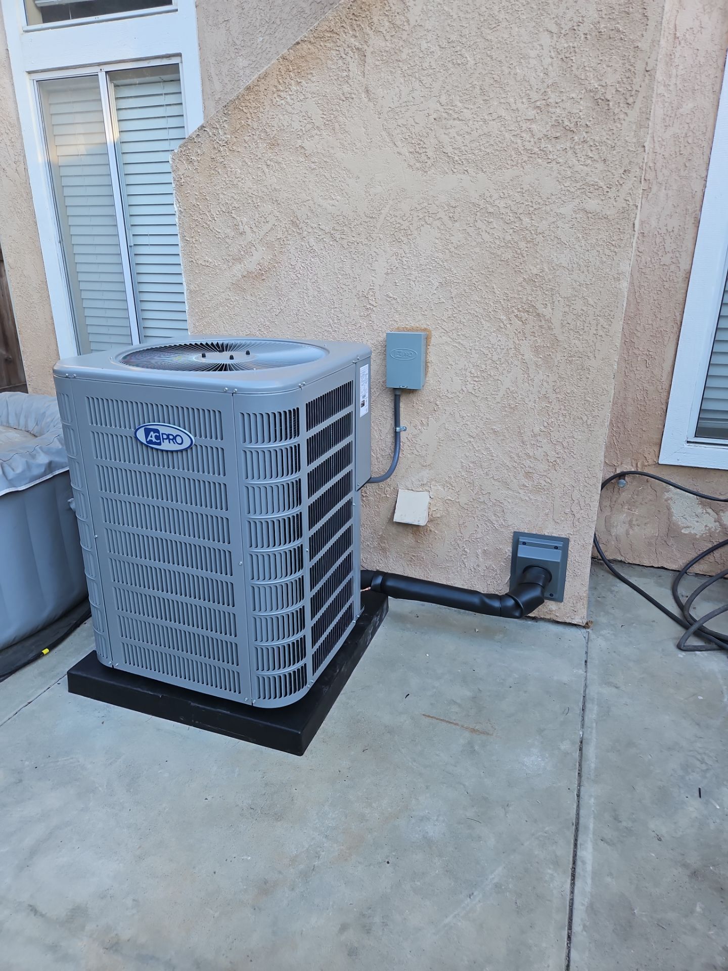 An outdoor air conditioning unit, gray and boxy, sits on a black platform next to a beige stucco wall.