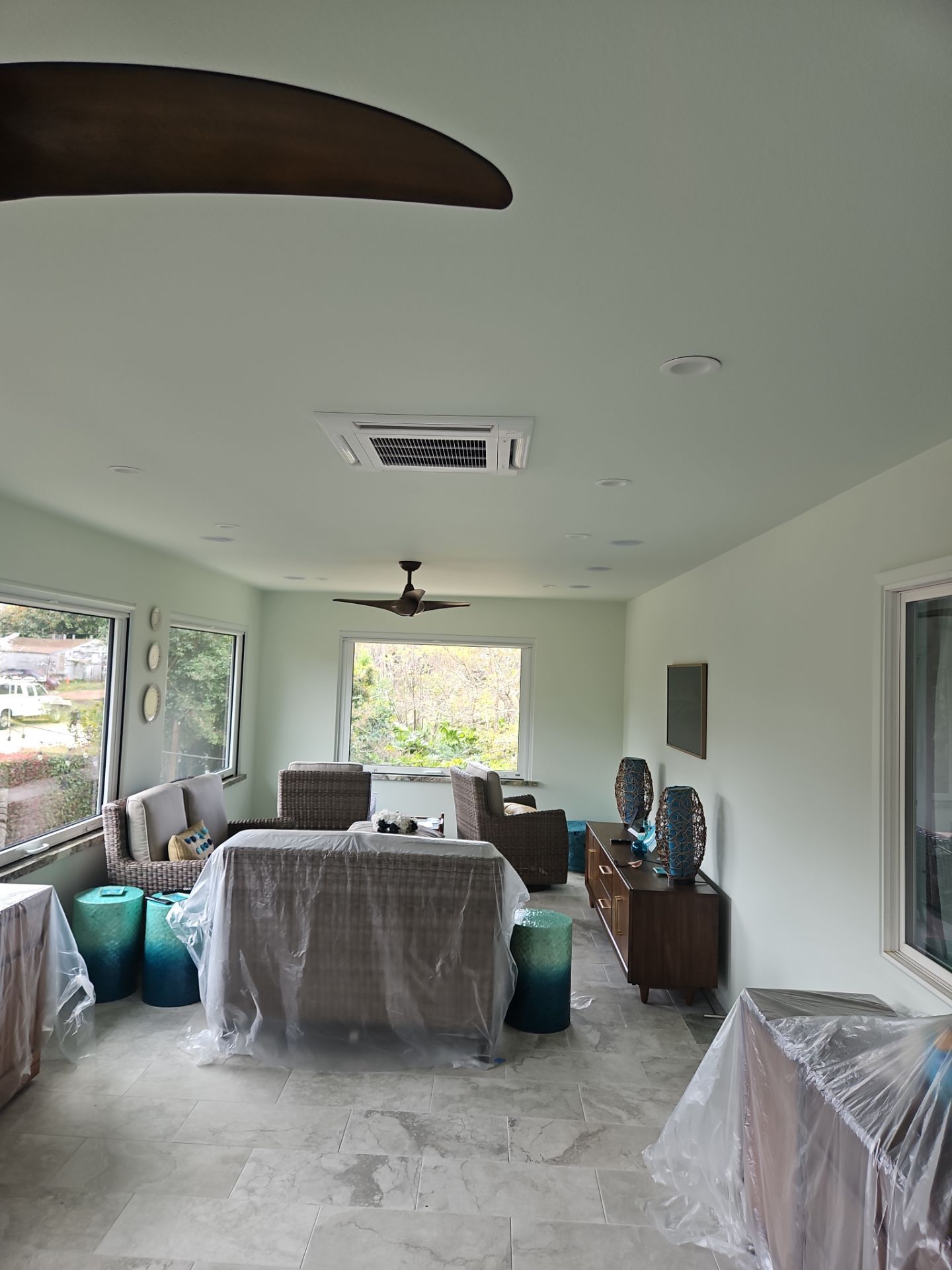 Interior of a light green room with furniture covered in plastic. There are windows, a ceiling fan, and decorative objects.