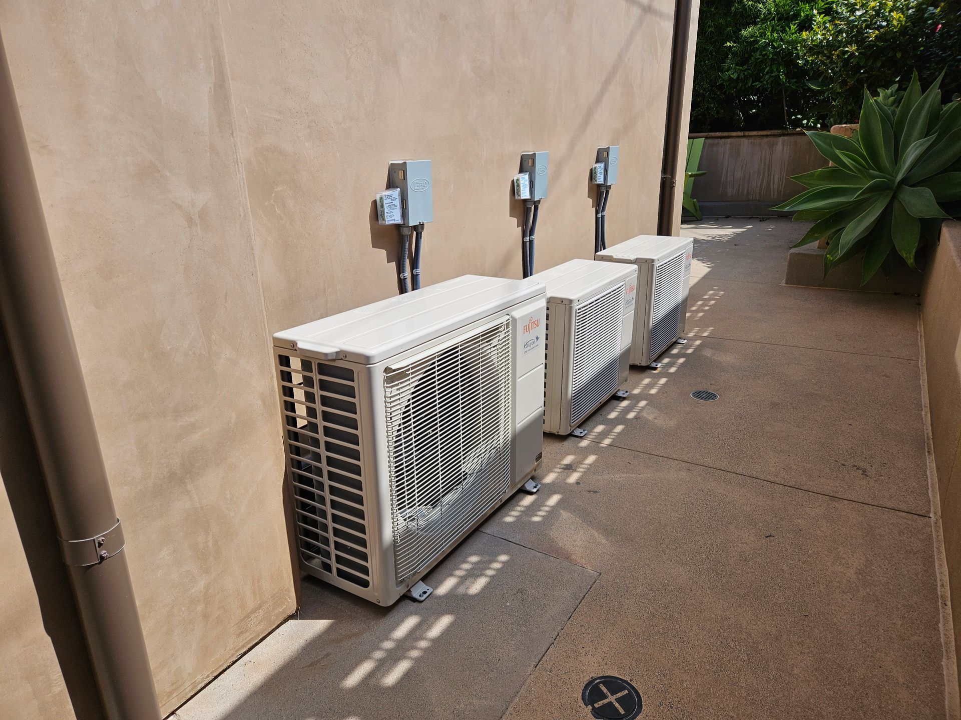 Three white air conditioning units against a beige wall with conduit and power boxes in an outdoor alley.