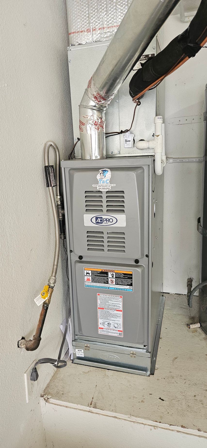 A gray HVAC furnace with a metal duct and gas pipe, indoors, on a concrete ledge.