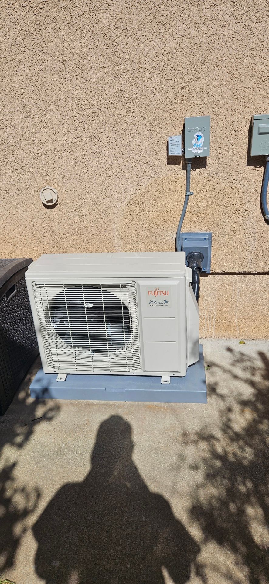 An AC unit is mounted on a concrete pad next to a textured beige wall. Electrical boxes are mounted above.