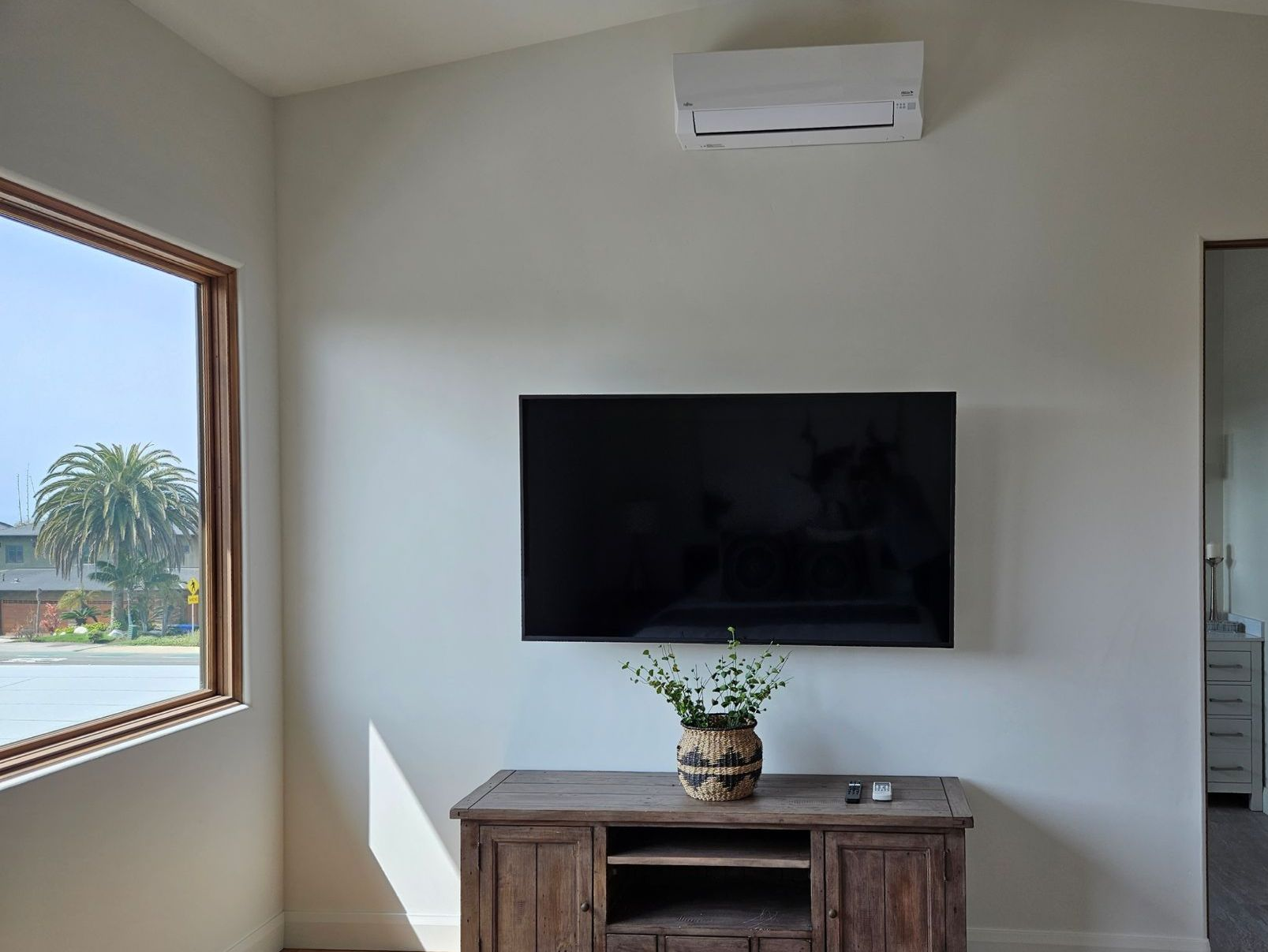 Living room with a wall-mounted TV, AC unit, and wooden cabinet. Window with palm tree view.