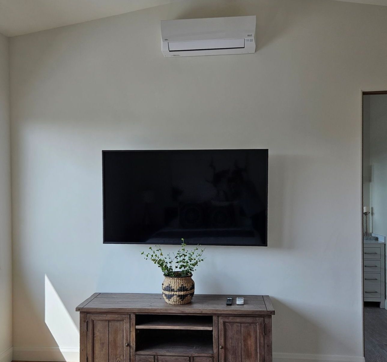A TV mounted on a white wall above a wooden cabinet with a vase of greenery; air conditioner above.