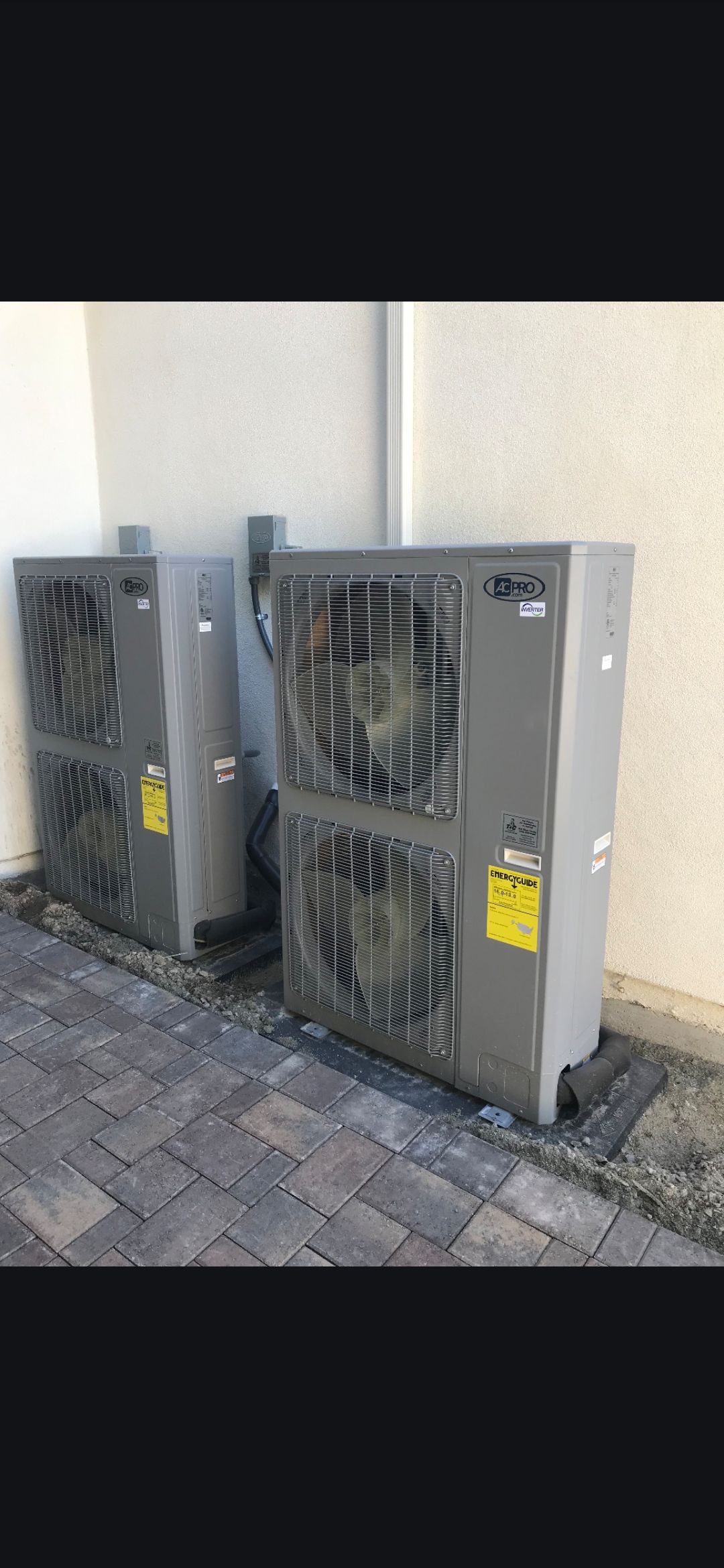 Three outdoor air conditioning units next to a light-colored wall on a stone surface.