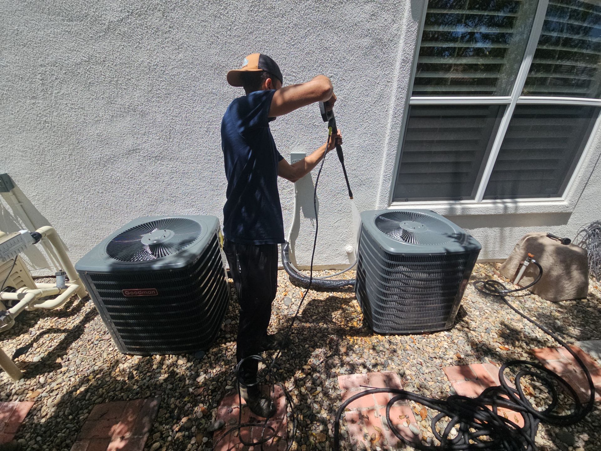 A person pressure washing two outdoor AC units on a gravel surface near a building with a window.