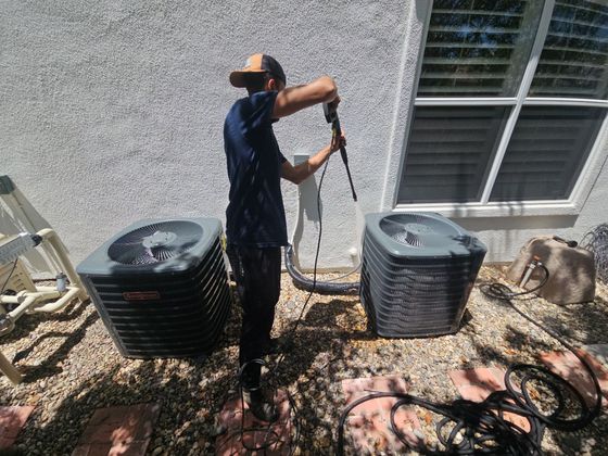 Man power washing two air conditioning units next to a window on a sunny day.