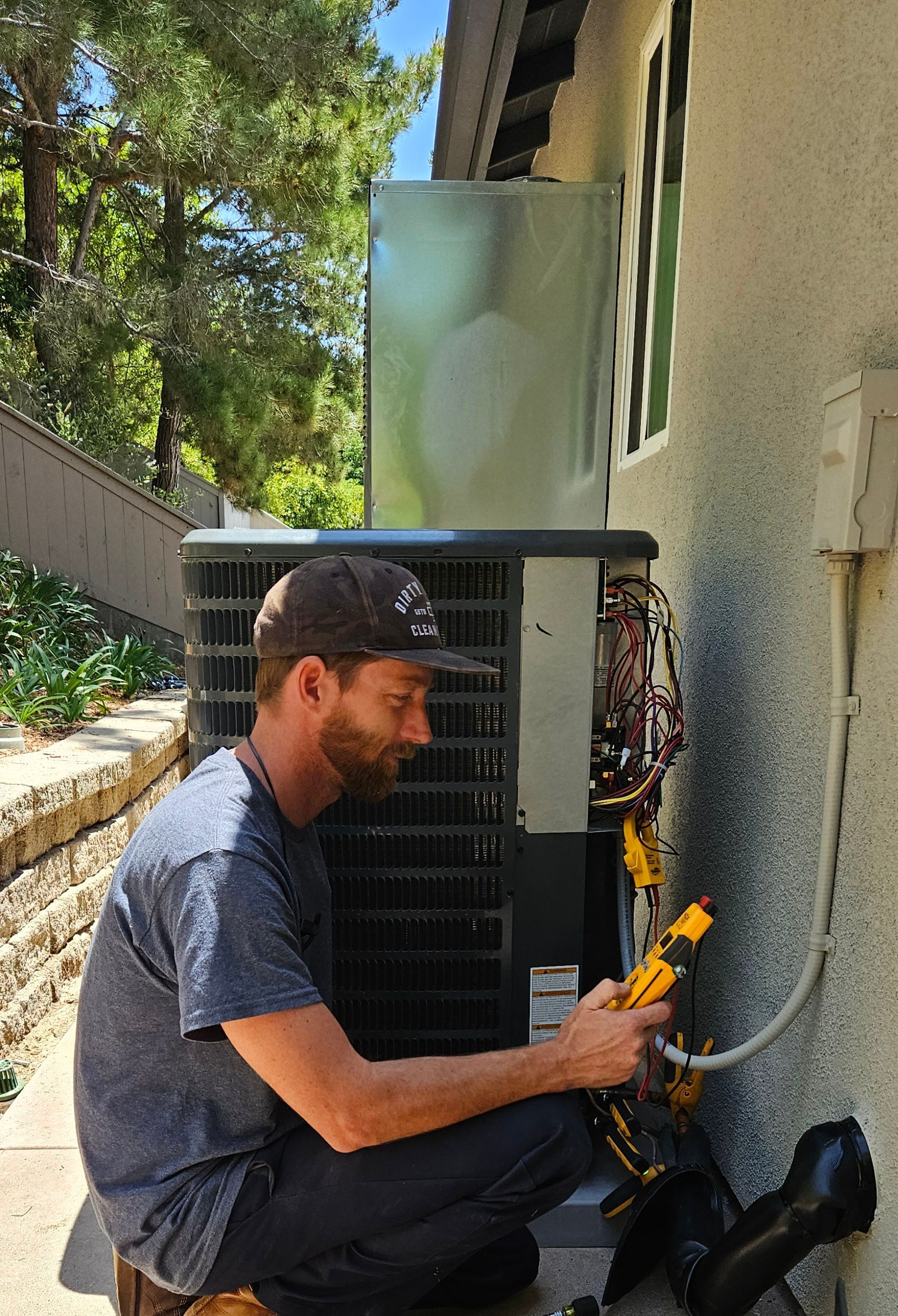 Man in cap repairing AC unit outside, holding a yellow tool.