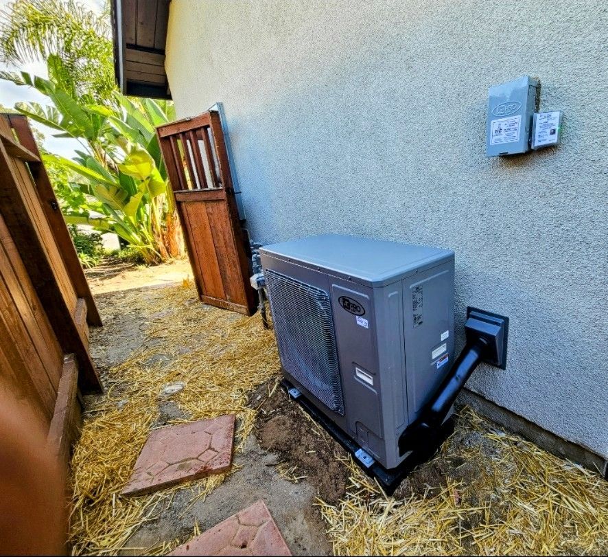 A gray pool heater installed outside a beige stucco wall, next to a wooden door and fence.