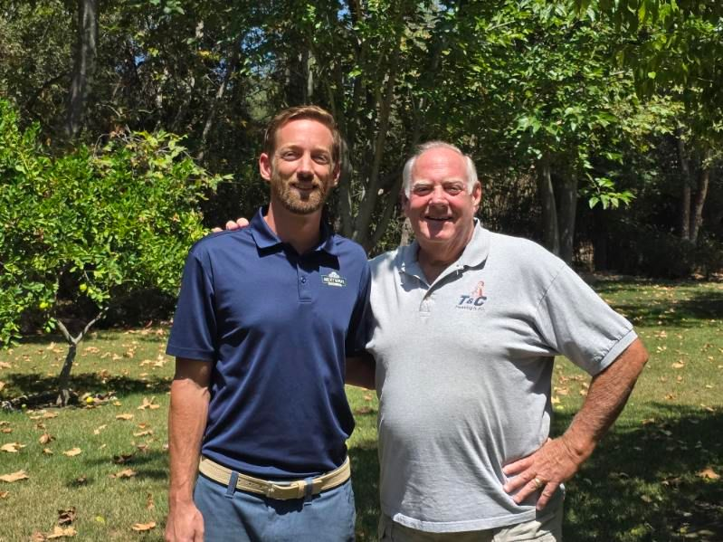 Two men, one in blue and one in gray, smile while standing in a yard with trees.