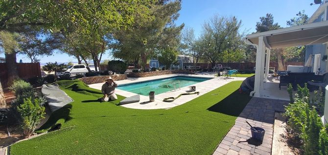 Backyard with pool and artificial grass installation; worker is kneeling. Blue sky.