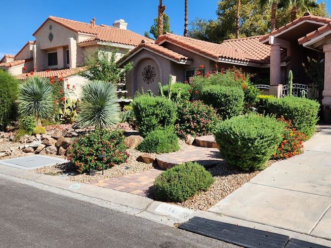 Residential landscaping with various green bushes, red flowers, and a red-tiled roof. Sunny day.