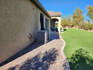 A brick pathway curves alongside a tan stucco house with a green lawn under a clear blue sky.