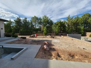 Backyard under construction: dirt, concrete, rocks, trees, blue sky, and a small construction vehicle.