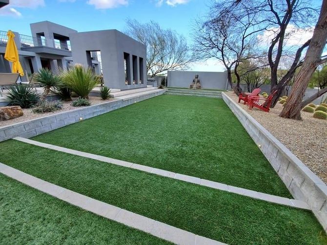 Artificial turf backyard with modern architecture, concrete borders, and two red chairs.