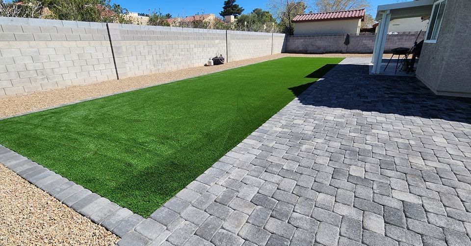 Backyard with green turf, stone patio, gravel, and a concrete wall.