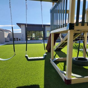 Playground with swings and slide in a grassy yard; modern house in the background on a sunny day.
