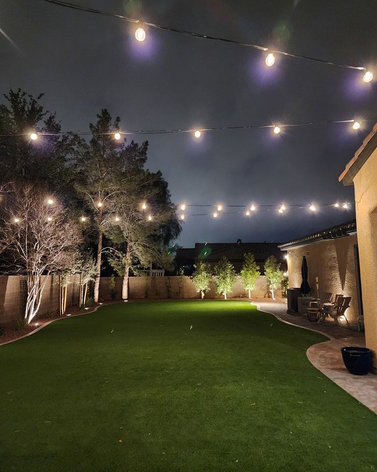 Nighttime backyard with string lights, trees, green grass, and illuminated house.