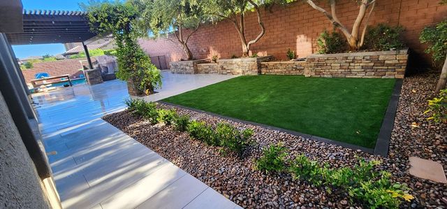 Backyard with green grass, gravel, plants, stone wall, and a patio.