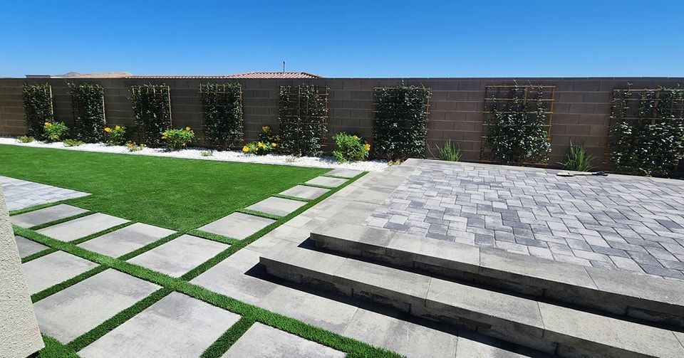 Backyard with green grass, stone patio, and planted wall with blue sky.