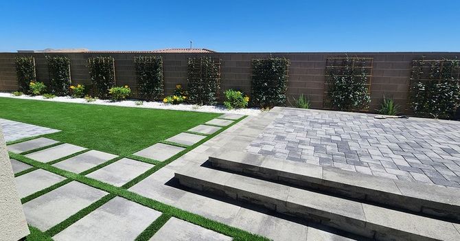 Backyard with green grass, stone patio, and planted wall with blue sky.