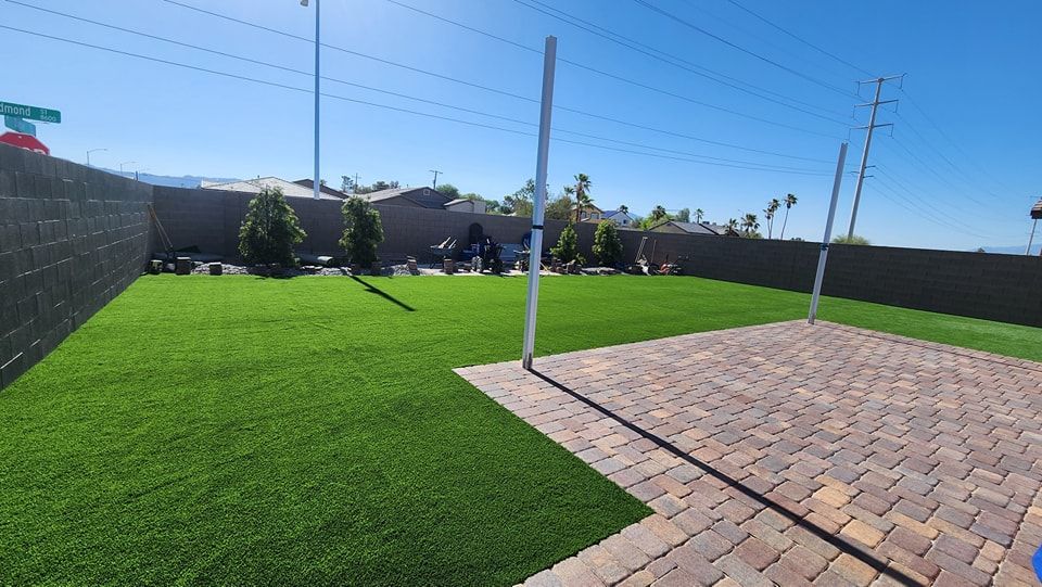 Backyard with artificial turf, brick patio, and a black fence under a blue sky.