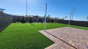 Backyard with artificial turf, brick patio, and a black fence under a blue sky.