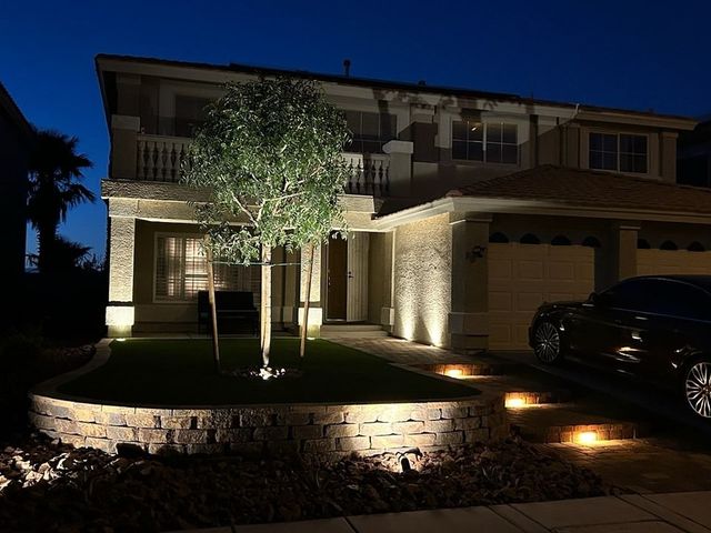 Lit-up house at night, with a tree and low-lying lights illuminating the yard and facade. A car is parked in the driveway.