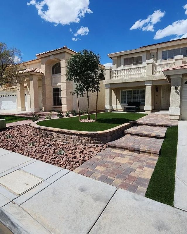 Two-story homes with landscaped yards and a brick pathway under a bright blue sky.