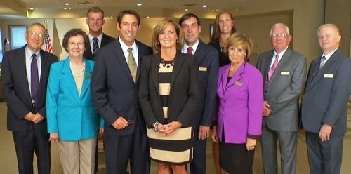 A group of nine people in professional business attire pose for a formal group portrait in a brightly lit indoor space.