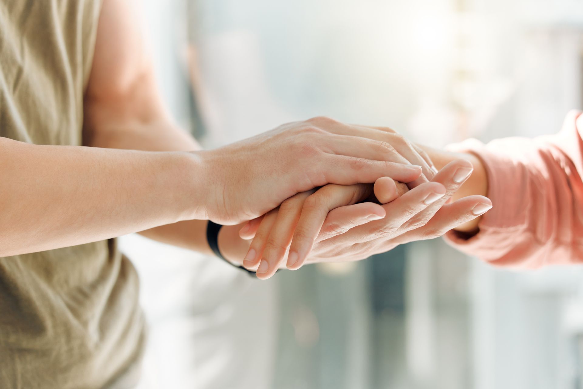 Two people hold hands in a gesture of support or comfort, with soft, bright natural lighting in the background.