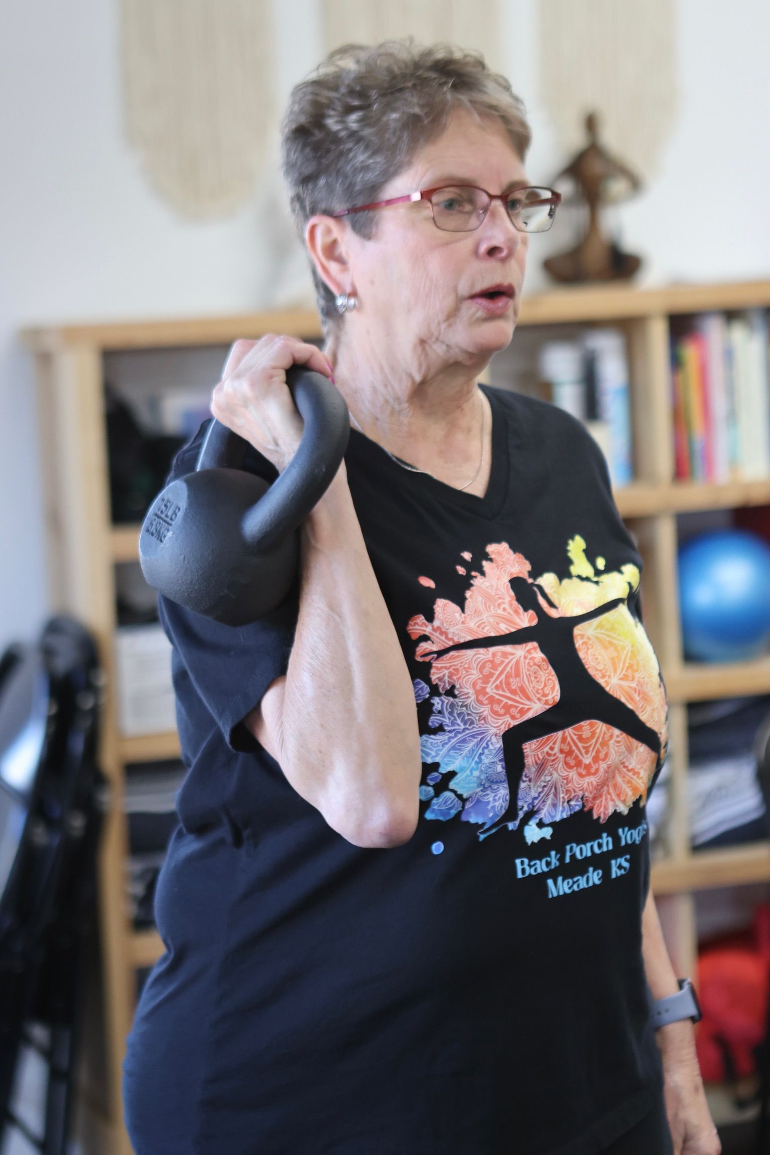 A woman wearing a black shirt with a basketball on it is lifting a kettlebell