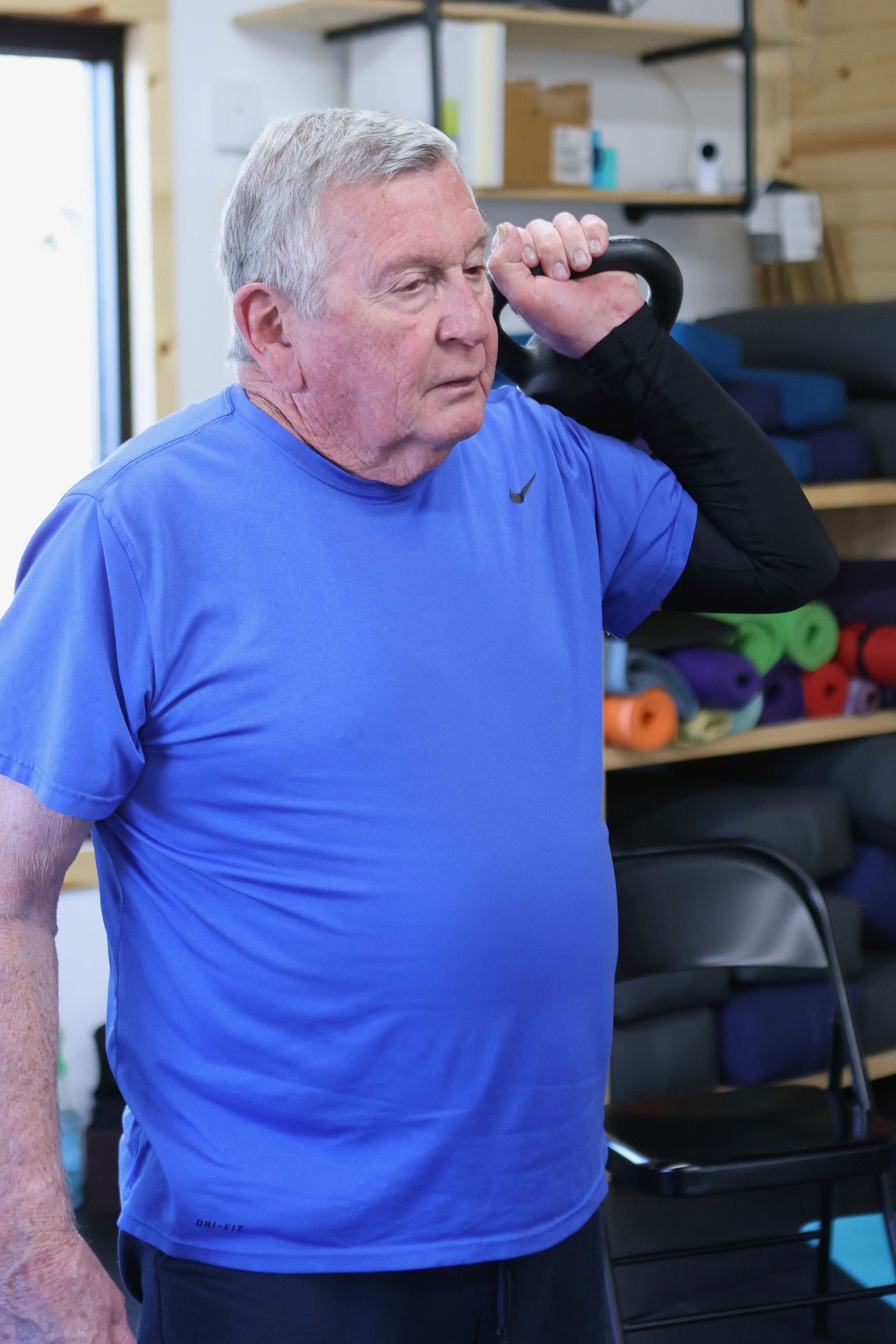 An older man in a blue shirt is lifting a kettlebell.