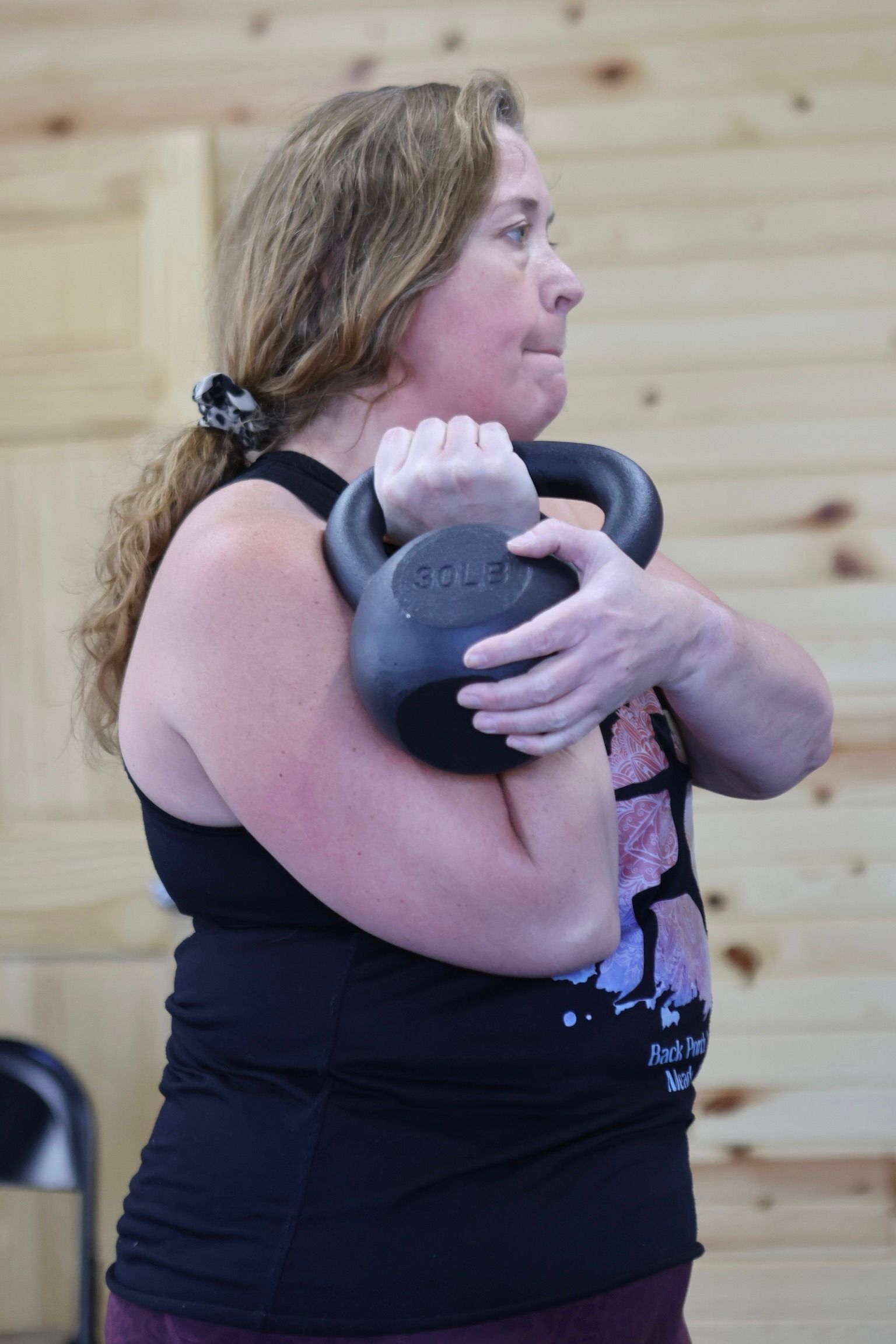 A woman is holding a kettlebell in her hands in front of a wooden wall.