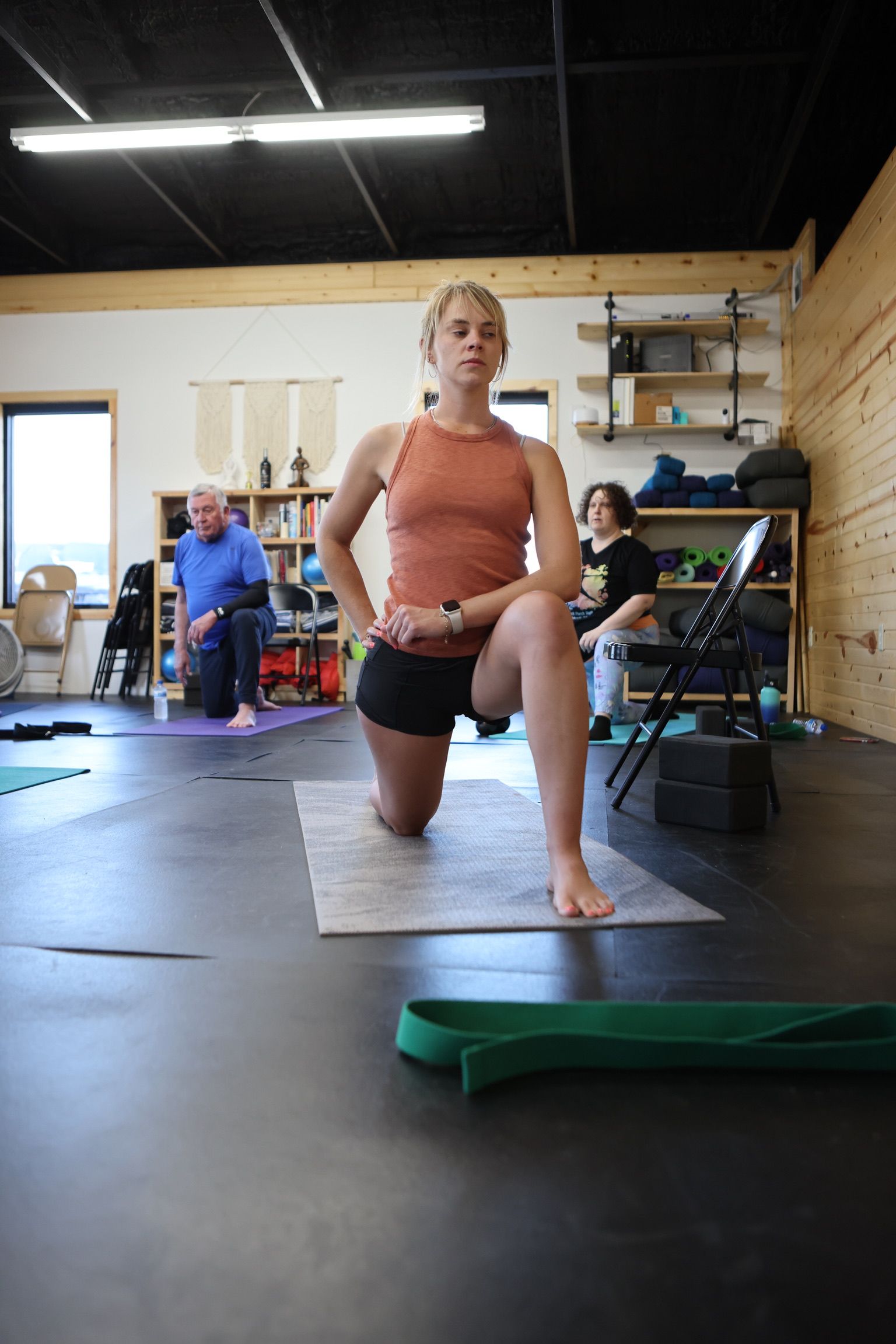 A woman is kneeling on a yoga mat in a gym.
