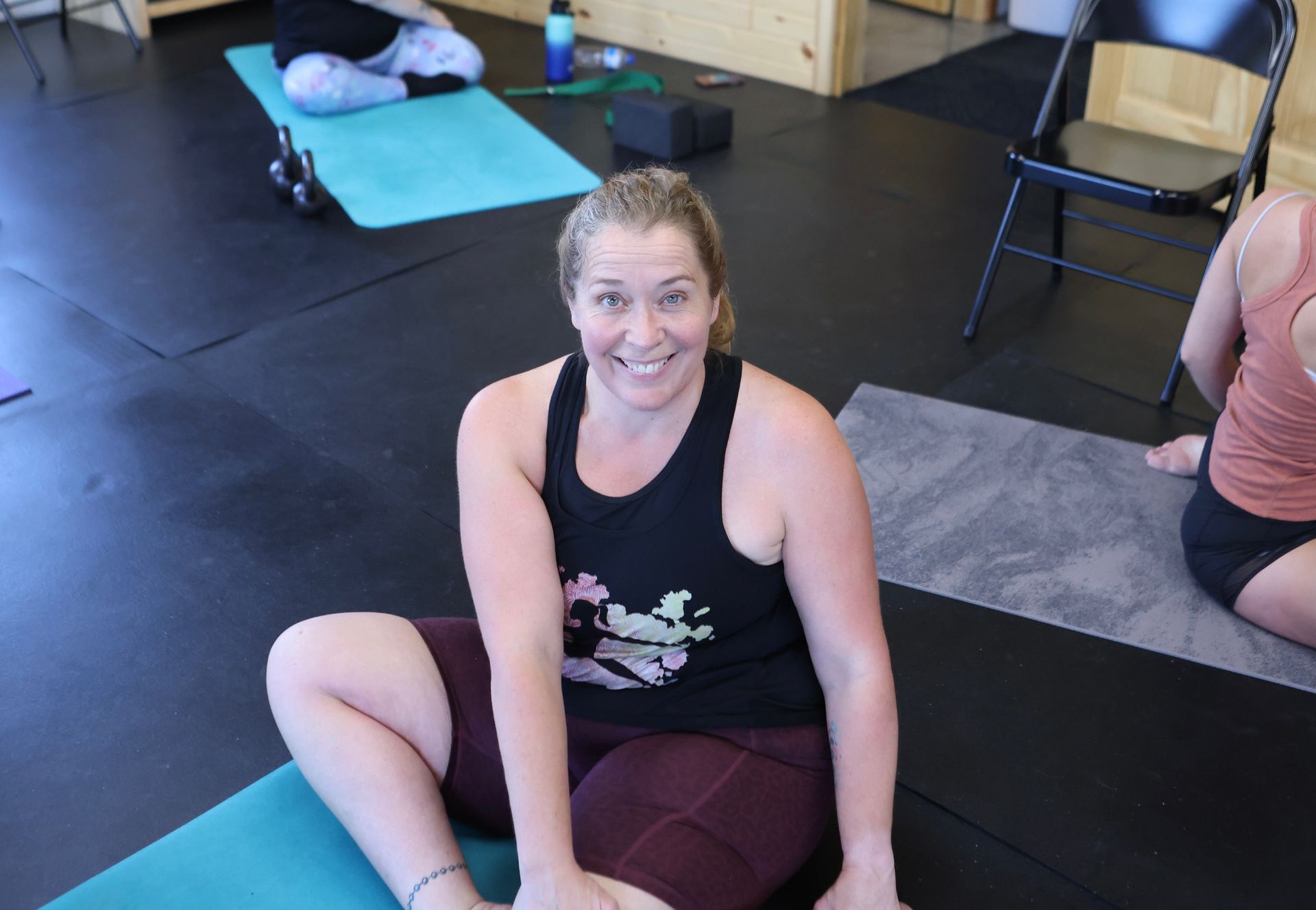 A woman is smiling while sitting on a yoga mat in a gym.