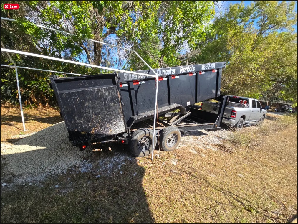 Red excavator loading debris into a black dumpster near a house.