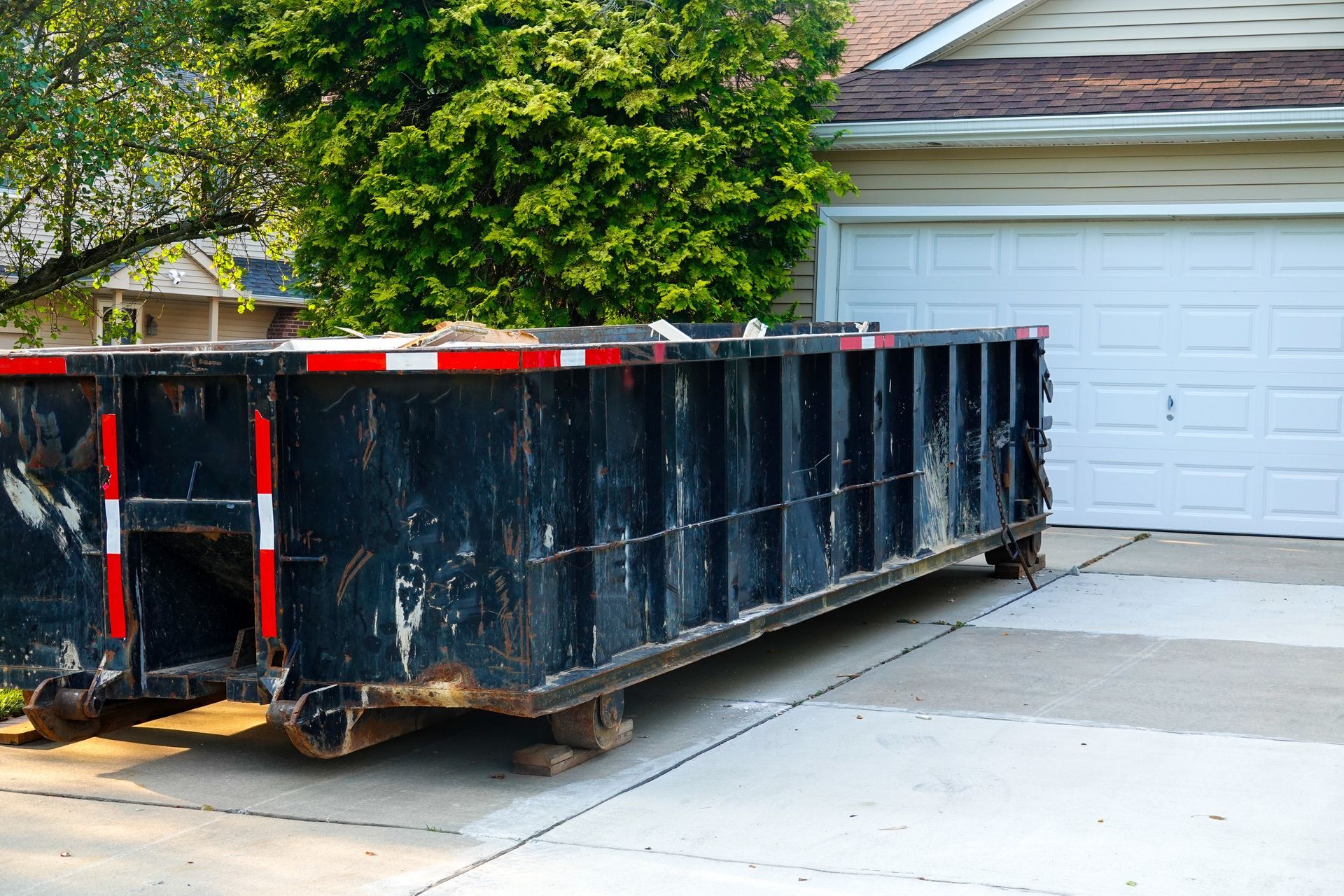Black dumpster on a driveway in front of a white garage door; a house and tree are in the background.