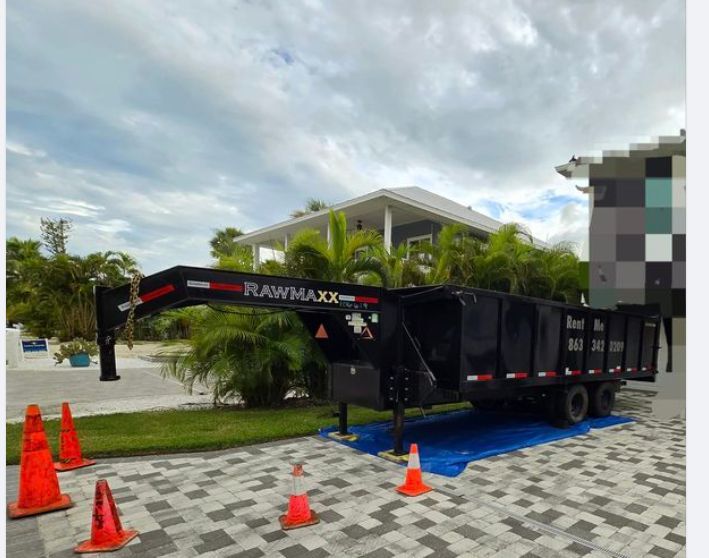 Black Rawmaxx dump trailer parked on a driveway with traffic cones, cloudy sky, and a house in the background.