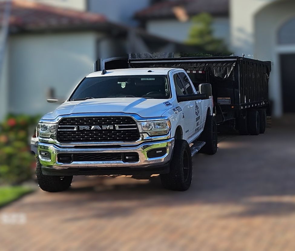 White Ram truck towing a black dump trailer parked on a driveway in front of a house.