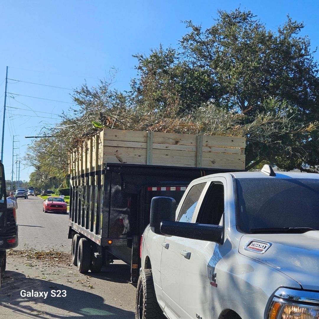 White pickup truck towing a black dump truck filled with tree branches on a sunny road.