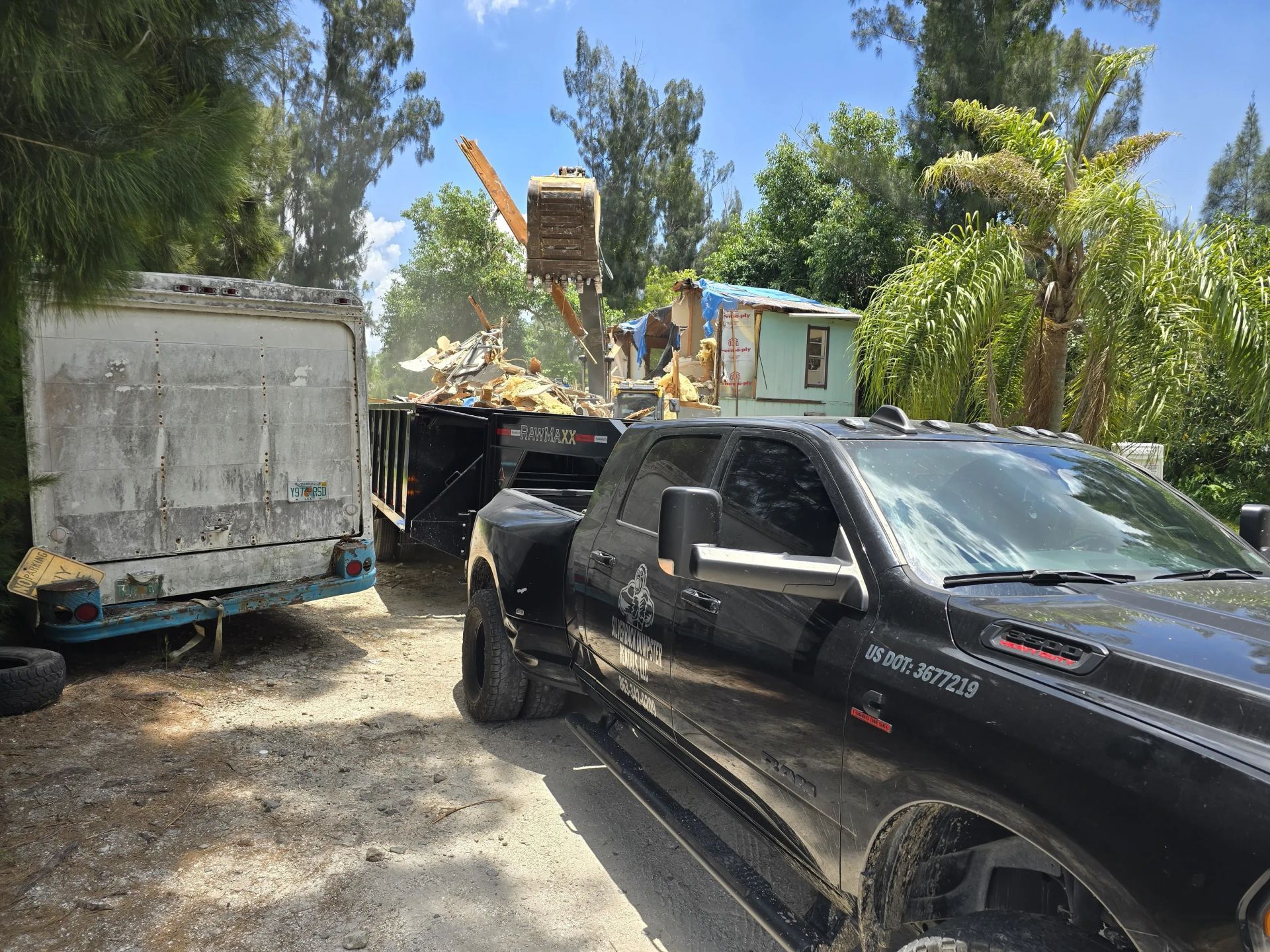 Black pickup truck hauling a trailer as an excavator loads debris. Outdoors, sunny day.