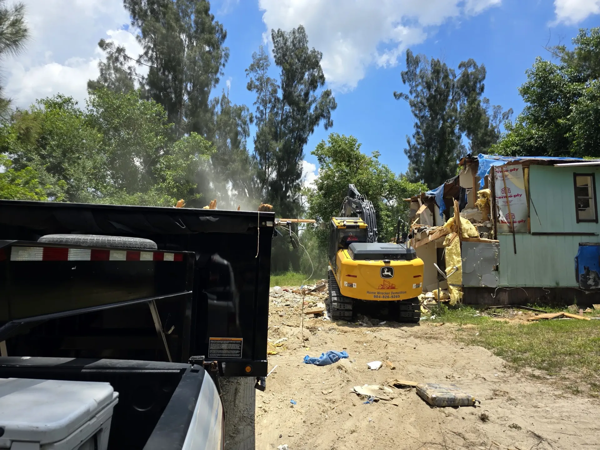Mini excavator demolishing a small structure, debris flying. Dump truck nearby on a sunny day.