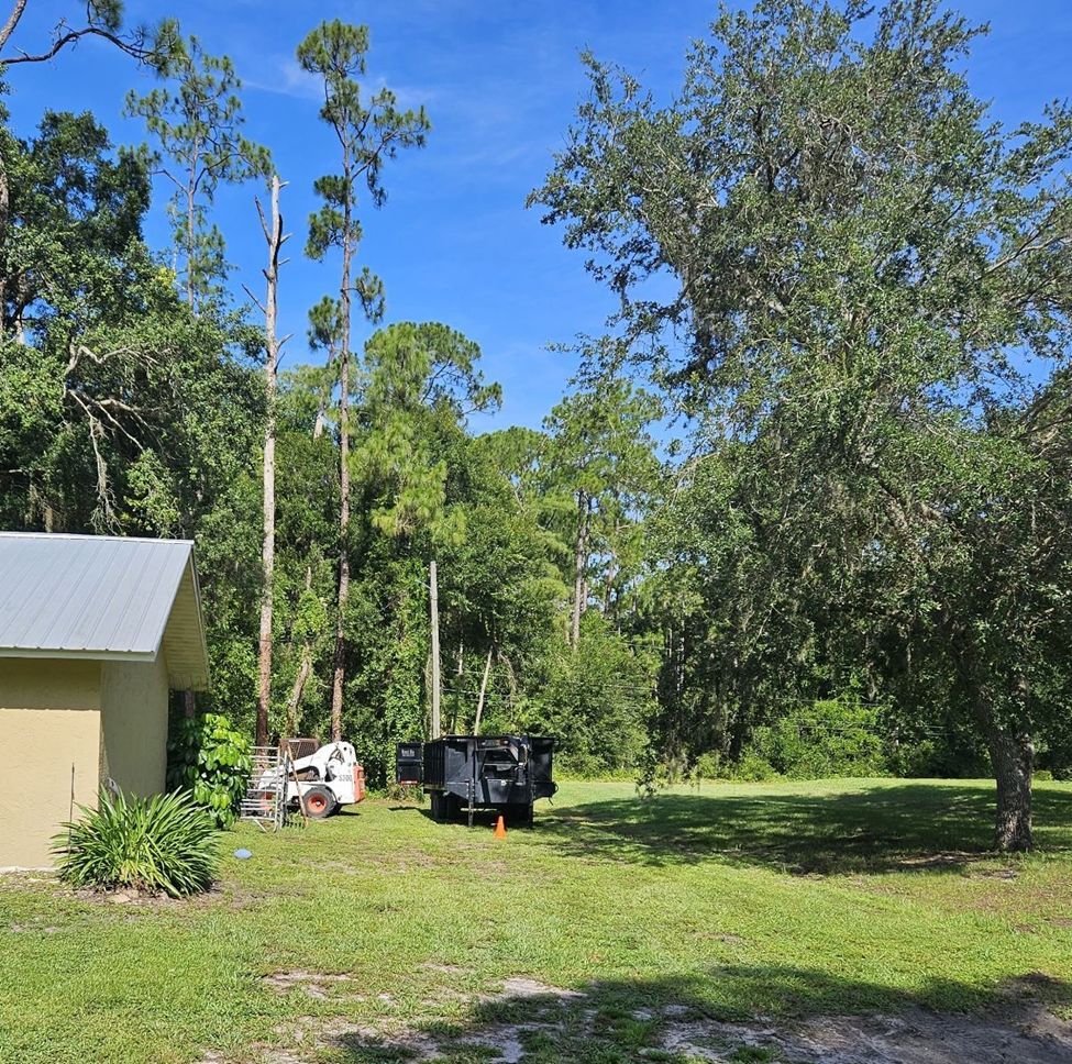 A yellow building next to a field. A tree stump, skid steer, and trailer in yard; blue sky.