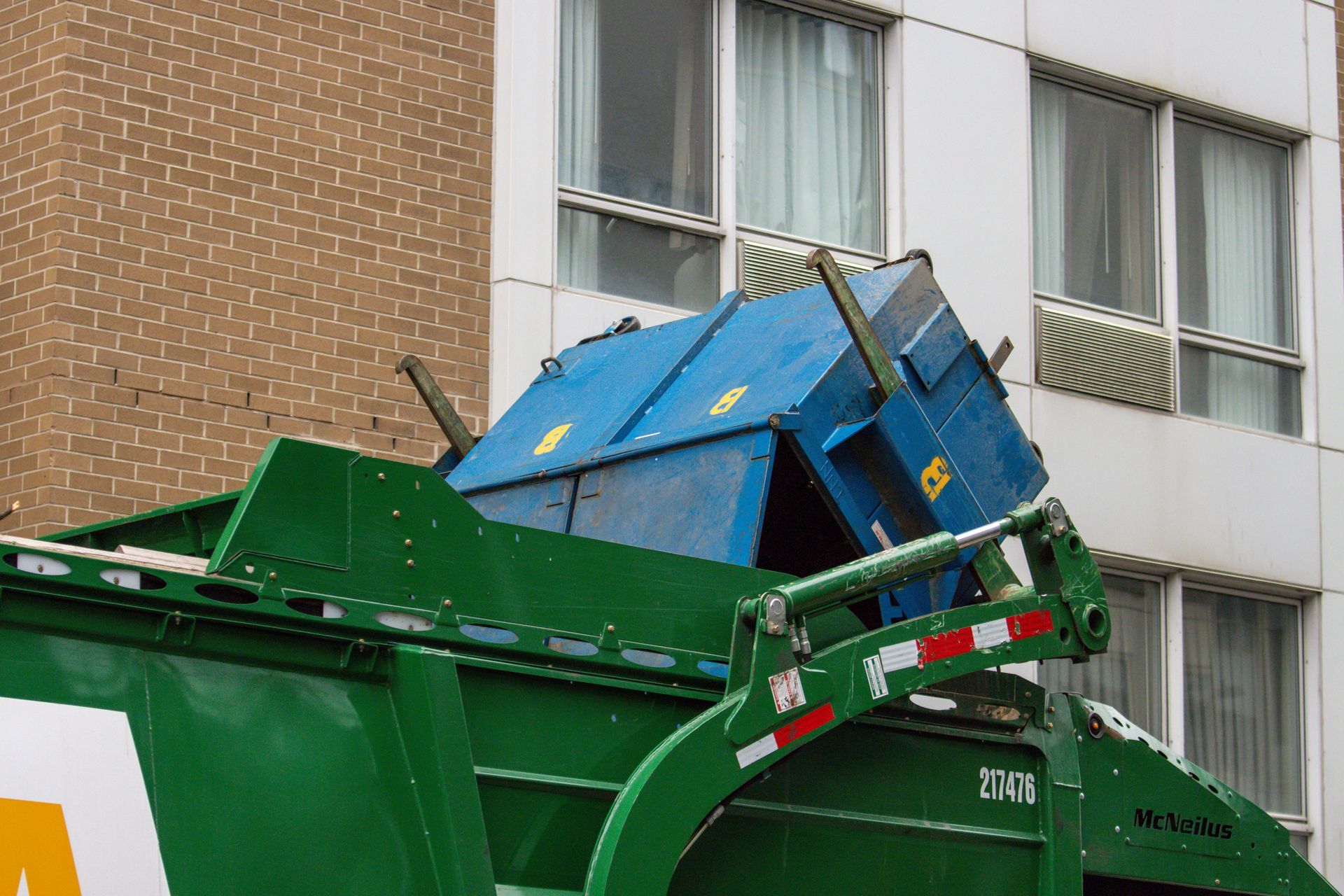 Green garbage truck lifting a blue dumpster. Building in the background.