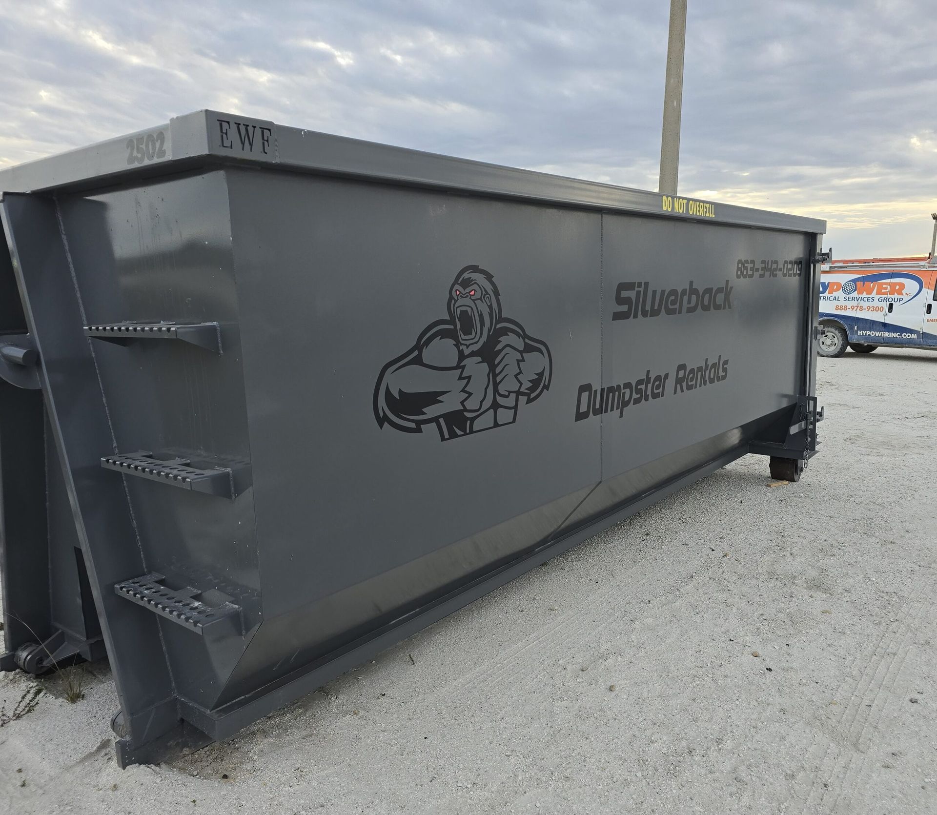 A blue dumpster being emptied by a green garbage truck in front of a brick building.